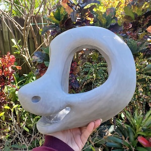 Op de afbeelding: Een lichtgrijze, abstracte sculptuur met een grote cirkelvormige opening. De sculptuur heeft een gladde, afgeronde vorm met een klein gaatje aan de zijkant. Het object wordt vastgehouden voor een achtergrond van groen en rood gebladerte.