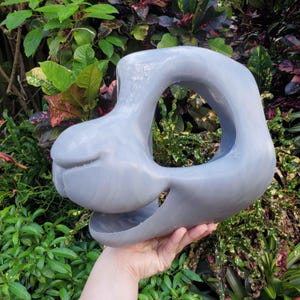 May include: A gray, abstract sculpture with a rounded, open center. The sculpture has a smooth, matte finish and is held in a hand against a backdrop of green foliage. The form has a unique, organic shape.