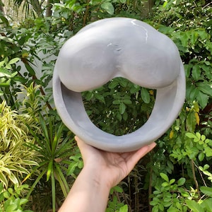 Può includere: Una scultura astratta in ceramica grigia con una forma arrotondata e una grande apertura. La scultura ha una superficie liscia ed è tenuta davanti a uno sfondo di fogliame verde.