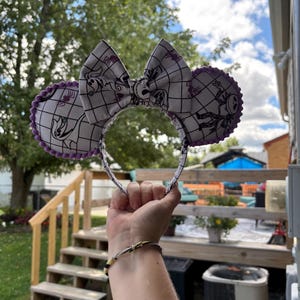 May include: A pair of mouse ears with a large bow and circular ear details. The ears and bow feature a black and white grid pattern with purple accents and Halloween-themed illustrations. The headband is held up against a background of greenery and a blue sky.