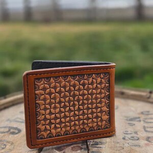 May include: A bi-fold wallet made of tan leather featuring a dark brown geometric floral pattern embossed on the front panel. The wallet has dark brown stitching along the edges and a smooth tan leather border framing the intricate stamped design.