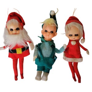 Puede incluir: Tres adornos navideños vintage. Uno es Papá Noel con un traje rojo con barba blanca y cinturón negro. El segundo es un elfo con un atuendo verde azulado. El tercero es una niña con un vestido rojo con adornos blancos.