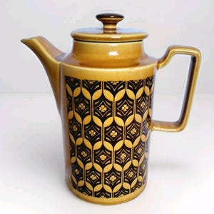 May include: A vintage ceramic coffee pot with a lid and handle. The pot is a warm tan color with a black and gold geometric pattern. The spout is on the left, and the handle is on the right. The lid has a small, dark brown knob.