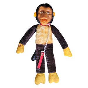 May include: A plush monkey toy with a moulded plastic face, black fur on the limbs and head, and a tan chest. The monkey features yellow hands and feet, a red collar with a silver metal chain, and a red strap attached to the chain hanging down.
