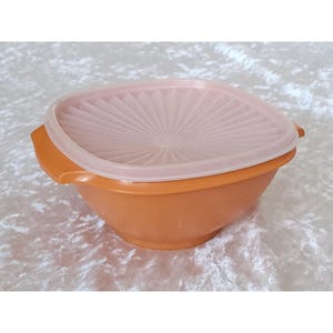 Puede incluir: Un cuenco de plástico naranja con una tapa a juego. La tapa es cuadrada con un diseño blanco en forma de abanico. El cuenco tiene dos asas a cada lado. El cuenco y la tapa están hechos de plástico duradero.