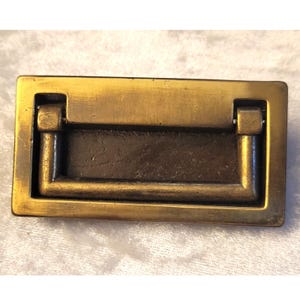 May include: A rectangular brass flush mount drawer pull featuring a recessed center and a hinged drop handle. The metal surface displays a brushed, aged finish with visible texture and dark patina within the recessed area of the hardware.