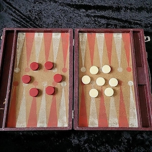 May include: Open backgammon board with red and white triangular markings. Red and white game pieces are arranged on the board. The board is housed in a burgundy case with a clasp.
