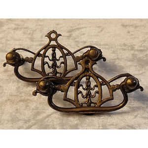 May include: Two matching brass drawer pulls featuring an ornate, openwork backplate with a central vertical motif and a curved, swinging handle. Each piece has a weathered, metallic finish and two circular mounting points for attachment.