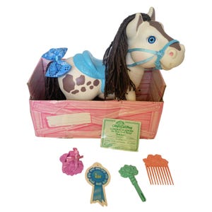 May include: A Crimp 'n Curl Pony toy named Midnight with a cream body, brown spots, and dark brown yarn mane. It wears a blue saddle and bridle with a blue bandana-print bow. Includes a pink basket, a 1st place ribbon, a green wand, and a comb.