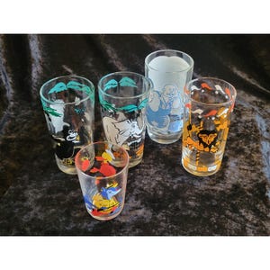 May include: A collection of five clear glass tumblers featuring colorful cartoon illustrations. The designs include a black gorilla, a white elephant, a blue circus performer, a yellow lion, and a small glass with a red and blue character graphic.