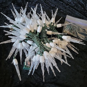 May include: A cluster of clear, icicle-shaped Christmas lights with white bases, strung on green wire. A small, clear plastic bag containing additional bulbs is visible. The lights are arranged on a dark surface.