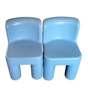 May include: Two light blue plastic chairs with rounded backrests and four sturdy cylindrical legs. Each chair features a molded seat and a smooth, matte finish. The brand name Little Tikes is embossed on the center of each backrest.