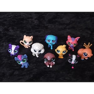 May include: A collection of colorful, small plastic animal figurines, including a cat, dog, skunk, and bird. The toys have large eyes and are set against a dark background. These collectible toys are popular with children.