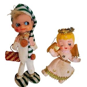 Op de afbeelding: Twee vintage-stijl beeldjes: een elf met een wit en groen gestreepte slaapmuts en pyjama, die een rood en wit gestreept cadeau vasthoudt, en een engel met blond haar, een gouden klatergoud halo, gouden vleugels en een witte jurk met gouden sterren.