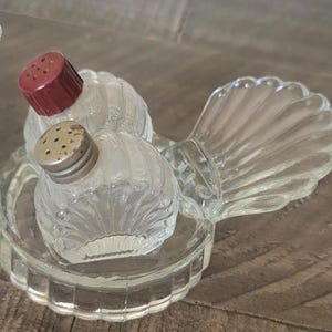 May include: Clear glass salt and pepper shakers with a shell-shaped design. The shakers have a ribbed texture and are topped with a red and silver metal lid. The set includes a clear glass tray with a scalloped edge.