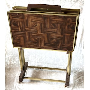May include: A set of four rectangular folding snack tables stored in a dark brown wooden rack with a handle. Each table features a dark brown wood grain surface with a geometric parquet pattern and a thin gold-toned metal frame border.