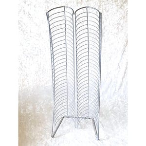 May include: A tall, silver-colored metal media storage rack featuring two vertical columns of curved wire slots designed to hold discs. The structure is made of thin, interconnected steel rods forming a series of parallel dividers for organization.