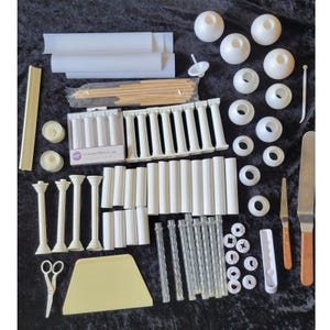 Puede incluir: Un conjunto de suministros para decorar pasteles que incluye pilares de plástico blanco, bases redondas, varillas transparentes texturizadas, tacos de madera, un raspador de plástico amarillo, espátulas de metal, tijeras y una caja etiquetada 4 Grecian Pillars.