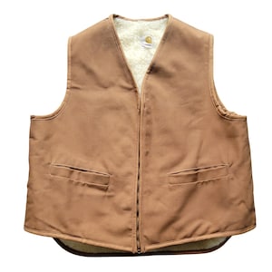 Puede incluir: Un chaleco Carhartt de color tostado con cierre de cremallera frontal completo, dos bolsillos horizontales ribeteados en la cintura y un forro grueso de forro polar sherpa color crema. La prenda tiene un cuello en V y un tejido exterior liso.