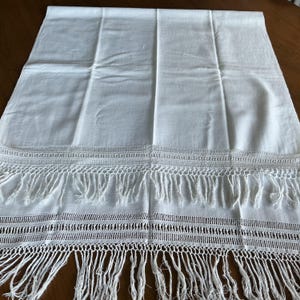 May include: A rectangular white textile runner featuring a smooth central panel, bordered by intricate horizontal bands of openwork crochet detailing, and finished with long, loose white fringe along the bottom edge of the fabric.