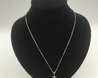 Op de afbeelding: Een zilveren ketting met een kleine kruisvormige hanger. Het kruis heeft een kleine, heldere steen in het midden. De ketting wordt getoond op een zwarte mannequin.