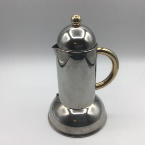 May include: A stainless steel stovetop espresso maker with a gold-colored handle and lid finial. The coffee pot has a cylindrical body and a rounded base. The lid and base are shiny, reflecting the light.