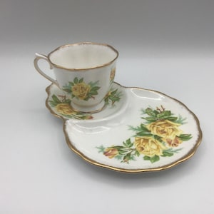 May include: A white teacup and saucer set with a yellow rose floral design and gold trim. The saucer has a unique shape with a section for a small treat. The teacup is on the saucer.