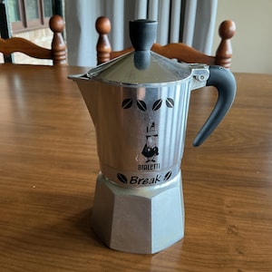 May include: A silver-coloured aluminium stovetop espresso maker with a black plastic handle and lid knob. The body features a black graphic of a person with a moustache, the text "BIALETTI" and "Break," and small leaf icons printed on the front.