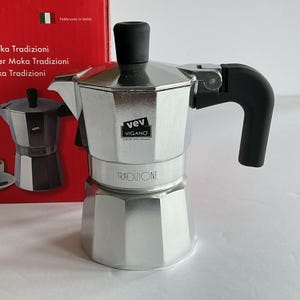 May include: A silver-colored stovetop espresso maker with a black handle and top. The words "vev VIGANO" and "TRADIZIONE" are printed on the espresso maker. The box behind the espresso maker has the Italian flag and the words "Fabbricato in Italia".