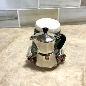 May include: A small silver espresso maker with a black handle and top, set on a base decorated with coffee beans. The espresso maker is in front of a white and green cup.
