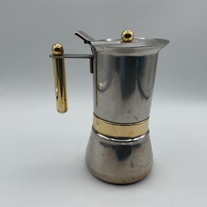 May include: Stainless steel stovetop espresso maker with a gold-colored handle and accents. The coffee maker has a silver body with a gold band around the middle and a gold knob on top. The handle is a gold color.