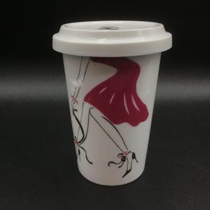 May include: White ceramic travel mug with a matching lid. The mug features a graphic of a woman in a red dress and high heels. The design is in black and red. The mug is cylindrical and has a white lid.