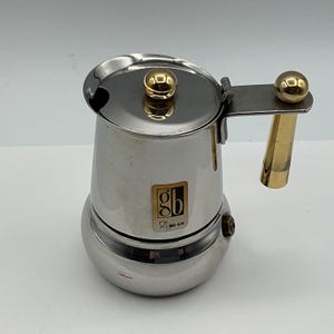 Peut inclure: Une cafetière italienne en acier inoxydable avec une finition polie. Elle présente un bouton sphérique doré sur le couvercle et une poignée dorée assortie. Un logo carré avec les lettres gb et le texte INOX 18/10 est centré sur le corps.