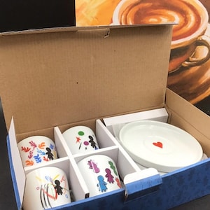 Peut inclure: Une boîte cadeau en carton contenant trois tasses en céramique blanche et une soucoupe blanche. Les tasses présentent des motifs colorés de personnages en bâtonnets et des formes. La soucoupe a un petit cœur rouge au centre.