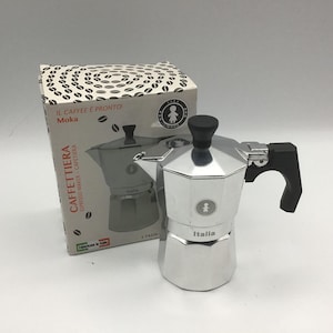 May include: A silver espresso maker with a black handle and top, next to a box with coffee bean illustrations and the text "IL CAFFEE È PRONTO! Moka CAFFETTIERA ESPRESSO MAKER - CAFETIERA". The espresso maker has the word "Italia" on it.