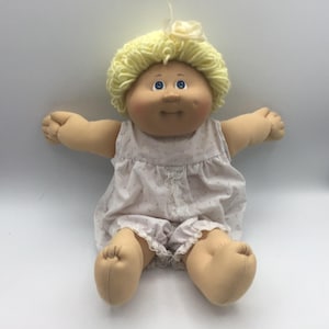 May include: A vintage doll with a soft, fabric body and a round, blushed face. The doll has blue eyes, a yellow yarn hairstyle, and is wearing a light pink romper with a floral pattern. The doll's arms and legs are outstretched.