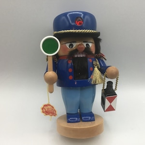 May include: A wooden nutcracker figurine of a guard in a blue uniform and hat, holding a green sign and a lantern. The figurine has a black mustache and beard, and is standing on a wooden base. The figurine is approximately 10 inches tall.