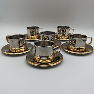 May include: A set of five metal cups and matching saucers. Each cup features a polished silver-tone upper body and a gold-tone base with a matching gold-tone handle. The saucers are circular with a gold-tone rim and a silver-tone centre.