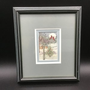 May include: Framed cross-stitch artwork depicting a winter scene with three red cardinals perched on a snow-covered tree branch. A small house and a pine tree are visible in the background.