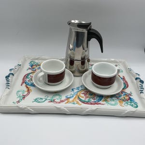 May include: A coffee set featuring a silver stovetop espresso maker, two small white espresso cups with brown bands, and saucers. The set rests on a rectangular tray with a white background and colorful floral designs.