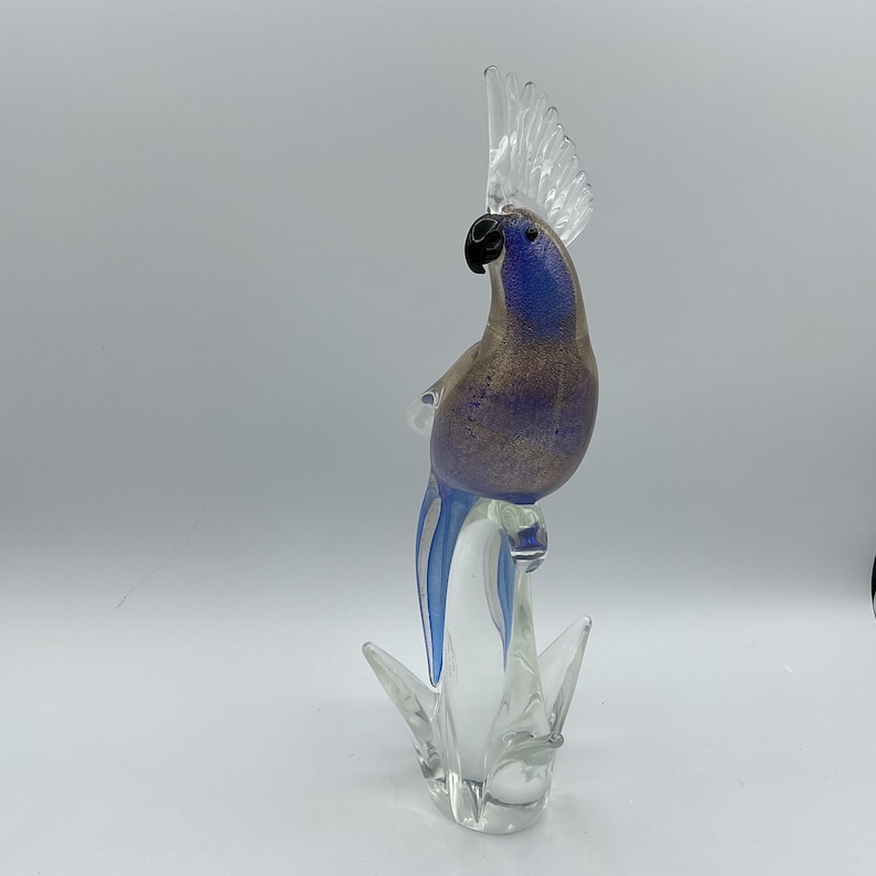 K&ouml;nnte beinhalten: Eine Glasskulptur eines Papageis mit blau-goldfarbenem K&ouml;rper, schwarzem Schnabel und einem durchsichtigen, f&auml;cherf&ouml;rmigen Kamm. Der Vogel sitzt auf einem Sockel aus klarem Glas mit spitzen, blattartigen Elementen.