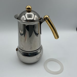 Könnte beinhalten: Ein Espressokocher aus Edelstahl mit goldfarbenem Griff und Akzenten. Der Kaffeezubereiter hat einen runden Boden und einen gewölbten Deckel. Eine weiße Gummidichtung liegt neben dem Kaffeezubereiter.