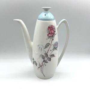 Peut inclure: Une cafetière en céramique blanche avec un couvercle et une poignée bleu clair. La cafetière est ornée d'une rose rose et de feuilles grises. Le bec verseur est long et fin.