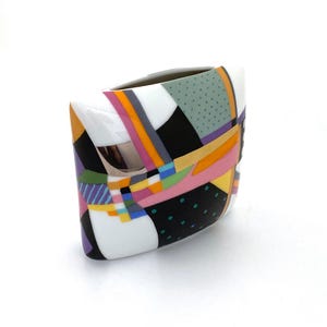 May include: A white ceramic vase with a geometric pattern in black, pink, yellow, green, blue, and purple. The vase has a rectangular shape with rounded corners.