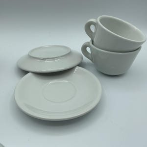 May include: A set of white ceramic teacups and saucers. Two cups are stacked, and two saucers are visible. The cups have rounded handles and a simple, classic design. The saucers are round with a slightly raised edge.