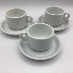 May include: Three white ceramic espresso cups with matching saucers. Each cup has a small handle and sits on a round saucer. The cups and saucers are all white and appear to be made of porcelain.
