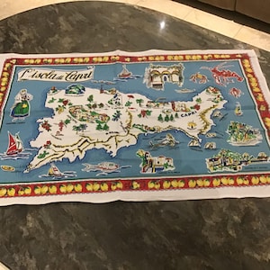 May include: A rectangular, white and blue Capri island map design on a fabric. The map features illustrations of boats, landmarks, and sea creatures. The border is red with yellow fruit illustrations and the text "L'Isola di Capri".
