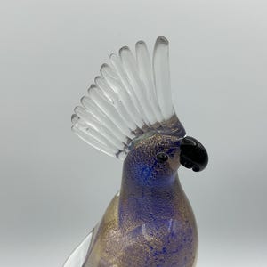 K&ouml;nnte beinhalten: Eine Glasskulptur eines Kakadus. Der Vogel hat einen klaren, f&auml;cherf&ouml;rmigen Kamm, einen schwarzen Schnabel und einen K&ouml;rper mit blauen und goldenen Flecken. Die Skulptur steht vor einem schlichten Hintergrund.