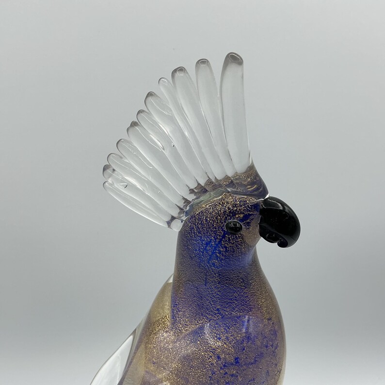 K&ouml;nnte beinhalten: Eine Glasskulptur eines Kakadus. Der Vogel hat einen klaren, f&auml;cherf&ouml;rmigen Kamm, einen schwarzen Schnabel und einen K&ouml;rper mit blauen und goldenen Flecken. Die Skulptur steht vor einem schlichten Hintergrund.