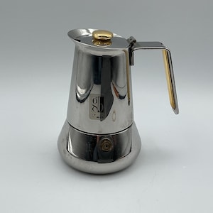 May include: A polished stainless steel stovetop espresso maker with a gold-toned knob on the lid and a gold-toned accent on the handle. The body features a square logo with the letters gb and a small symbol printed on the front of the upper chamber.
